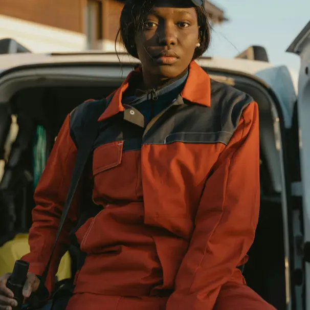 A worker in an orange and black uniform sitting outdoors near a vehicle.