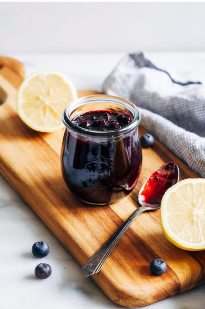 A jar of blueberry jam with lemon halves and a spoon on a wooden board.