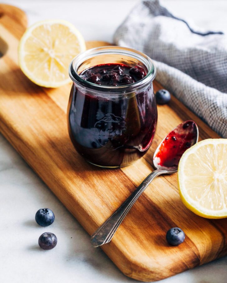 A jar of blueberry jam with lemon halves and a spoon on a wooden board.